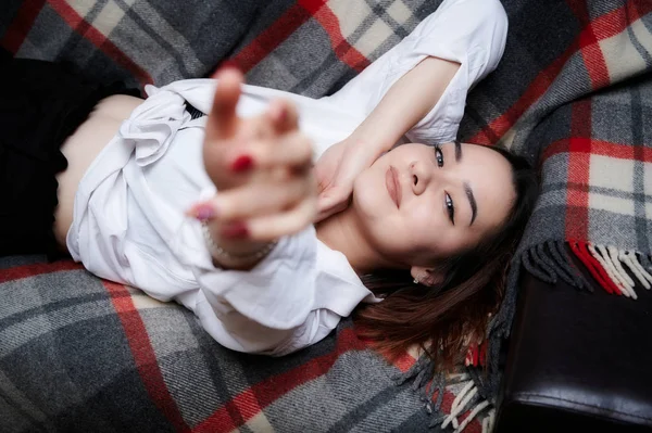 Portrait of a lovely brunette caucasian girl in a white shirt at home on the sofa. Lies and relaxes on a plaid in front of the camera in various poses. — Stock Photo, Image