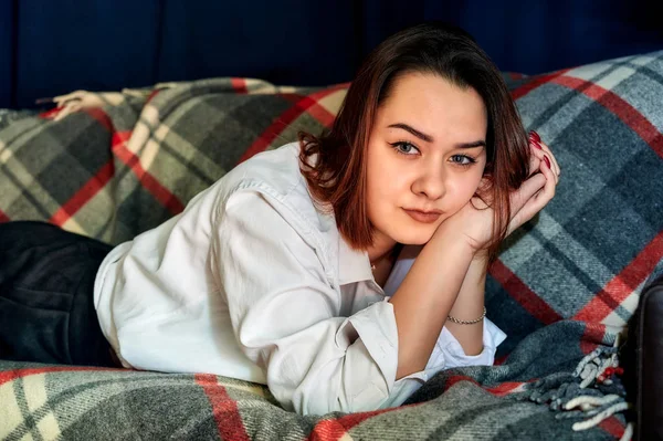 Portrait of a lovely brunette caucasian girl in a white shirt at home on the sofa. Lies and relaxes on a plaid in front of the camera in various poses. — Stock Photo, Image