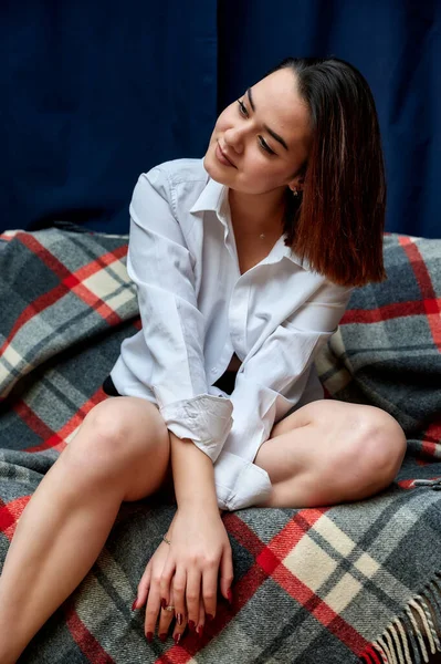 Portrait of a lovely brunette caucasian girl in a white shirt at home on the sofa. Lies and relaxes on a plaid in front of the camera in various poses. — 스톡 사진
