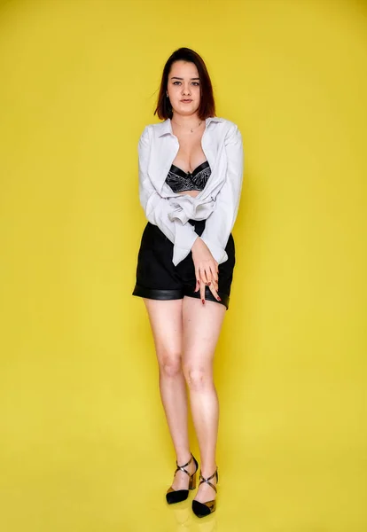 Full-length portrait of a pretty smiling brunette Caucasian girl in a white shirt and black shorts on a yellow background. Standing right in front of the camera in various poses. — 스톡 사진