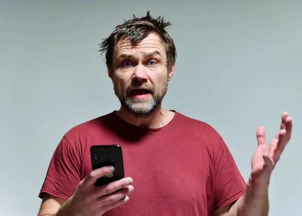 Retrato de um homem idoso selvagem e sem barba de 40 anos com um telefone em uma camiseta borgonha em um fundo cinza. Ele fica bem na frente da câmera, falando, mostrando emoções . — Fotografia de Stock