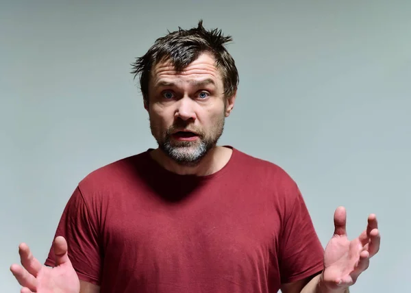 Portret van een wilde ongeschoren man van 40 jaar in een bordeaux t-shirt op een grijze achtergrond. Hij staat recht voor de camera, praat, toont emoties. Zwaait met zijn handen. — Stockfoto