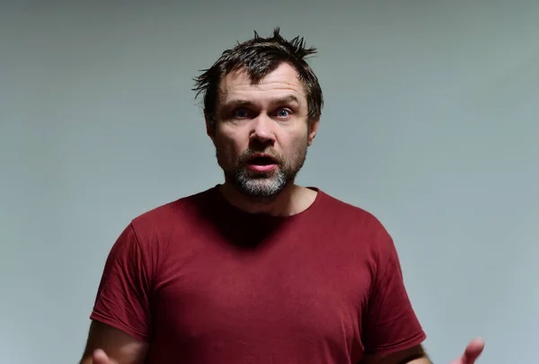 Portrait of a wild unkempt unshaven middle-aged man of 40 years in a burgundy t-shirt on a gray background. He stands right in front of the camera, talking, showing emotions. Waves his hands. — ストック写真