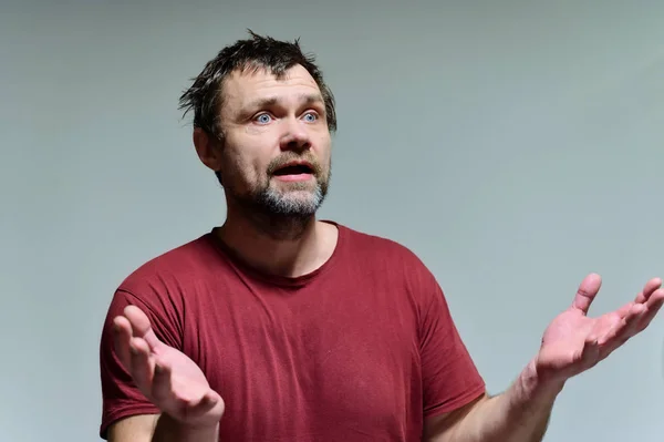 Portret van een wilde ongeschoren man van 40 jaar in een bordeaux t-shirt op een grijze achtergrond. Hij staat recht voor de camera, praat, toont emoties. Zwaait met zijn handen. — Stockfoto