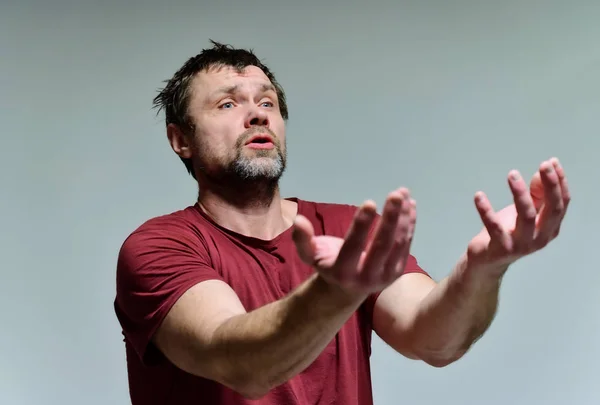 Portrait of a wild unkempt unshaven middle-aged man of 40 years in a burgundy t-shirt on a gray background. He stands right in front of the camera, talking, showing emotions. Waves his hands. — ストック写真