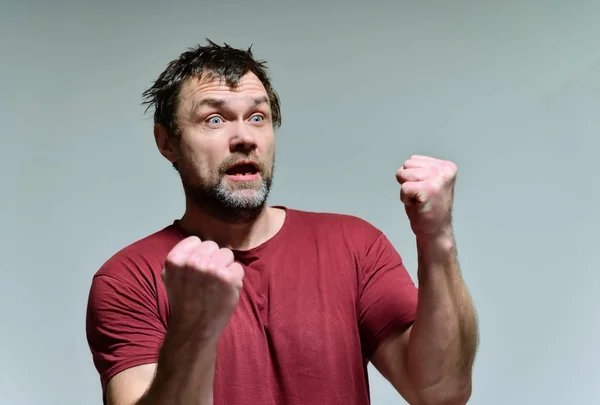 Portret van een wilde ongeschoren man van 40 jaar in een bordeaux t-shirt op een grijze achtergrond. Hij staat recht voor de camera, praat, toont emoties. Zwaait met zijn handen. — Stockfoto