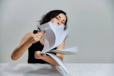 Beyaz arka planda siyah elbiseli, gülümseyen, konuşan esmer bir kadının portresi. Elinde bir klasörle kameranın önündeki masada canlı duygularla oturuyor..