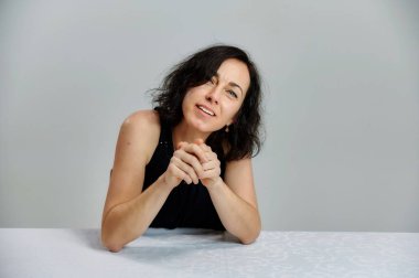 Model, kameranın önünde canlı duygularla bir masada oturur. Beyaz arka planda siyah elbiseli, gülümseyen, konuşan esmer bir kadının portresi..