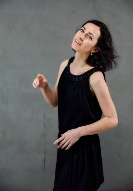 Model, canlı duygularla kameranın önünde duruyor. Gri arka planda siyah elbiseli, tatlı, gülümseyen esmer bir kadının dikey portresi..