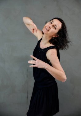Model, canlı duygularla kameranın önünde duruyor. Gri arka planda siyah elbiseli, tatlı, gülümseyen esmer bir kadının dikey portresi..