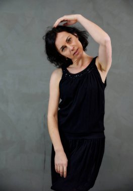 Model, canlı duygularla kameranın önünde duruyor. Gri arka planda siyah elbiseli, tatlı, gülümseyen esmer bir kadının dikey portresi..