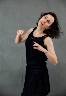 Model, canlı duygularla kameranın önünde duruyor. Gri arka planda siyah elbiseli, tatlı, gülümseyen esmer bir kadının dikey portresi..