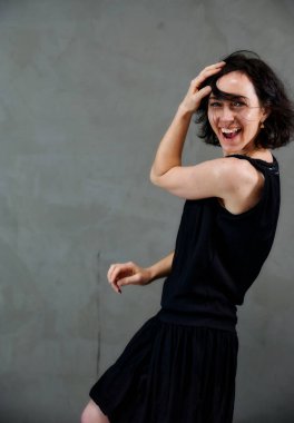 Model, canlı duygularla kameranın önünde duruyor. Gri arka planda siyah elbiseli, tatlı, gülümseyen esmer bir kadının dikey portresi..