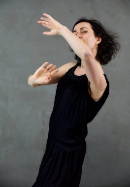 Model, canlı duygularla kameranın önünde duruyor. Gri arka planda siyah elbiseli, tatlı, gülümseyen esmer bir kadının dikey portresi..