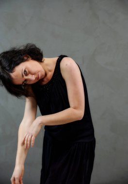 Model, canlı duygularla kameranın önünde duruyor. Gri arka planda siyah elbiseli, tatlı, gülümseyen esmer bir kadının dikey portresi..