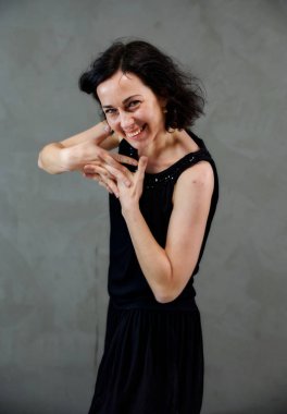 Model, canlı duygularla kameranın önünde duruyor. Gri arka planda siyah elbiseli, tatlı, gülümseyen esmer bir kadının dikey portresi..