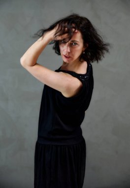 Model, canlı duygularla kameranın önünde duruyor. Gri arka planda siyah elbiseli, tatlı, gülümseyen esmer bir kadının dikey portresi..