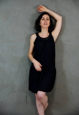 Model, canlı duygularla kameranın önünde duruyor. Gri arka planda siyah elbiseli, tatlı, gülümseyen esmer bir kadının dikey portresi..