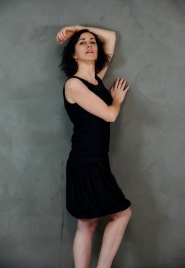 Model, canlı duygularla kameranın önünde duruyor. Gri arka planda siyah elbiseli, tatlı, gülümseyen esmer bir kadının dikey portresi..