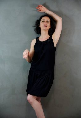 Model, canlı duygularla kameranın önünde duruyor. Gri arka planda siyah elbiseli, tatlı, gülümseyen esmer bir kadının dikey portresi..