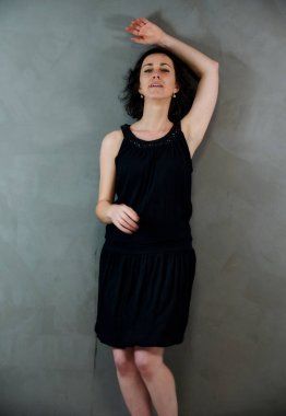 Model, canlı duygularla kameranın önünde duruyor. Gri arka planda siyah elbiseli, tatlı, gülümseyen esmer bir kadının dikey portresi..
