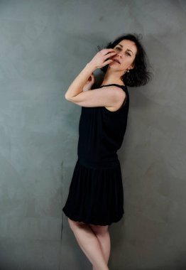 Model, canlı duygularla kameranın önünde duruyor. Gri arka planda siyah elbiseli, tatlı, gülümseyen esmer bir kadının dikey portresi..
