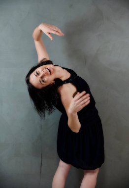 Model, canlı duygularla kameranın önünde duruyor. Gri arka planda siyah elbiseli, tatlı, gülümseyen esmer bir kadının dikey portresi..