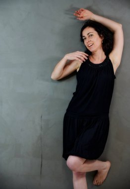 Model, canlı duygularla kameranın önünde duruyor. Gri arka planda siyah elbiseli, tatlı, gülümseyen esmer bir kadının dikey portresi..