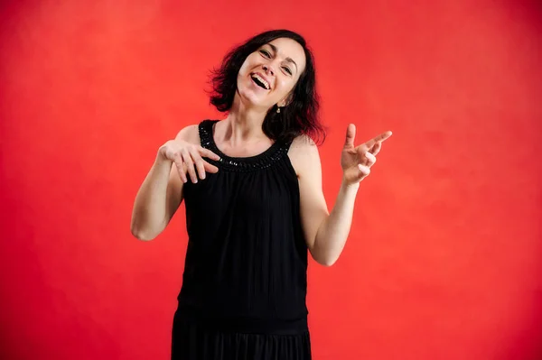 Retrato de uma mulher morena bonita em um vestido preto em um fundo vermelho. Mostra emoções vívidas com as mãos em diferentes poses bem na frente da câmera . — Fotografia de Stock