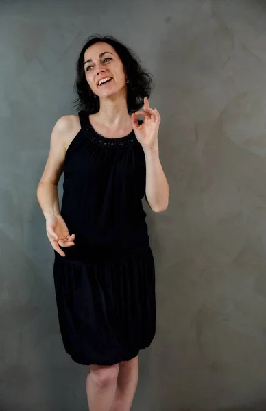 Retrato de larga duración de una mujer morena bastante emocional en vestido negro sobre fondo gris alternativo en el interior vintage. De pie justo delante de la cámara . —  Fotos de Stock