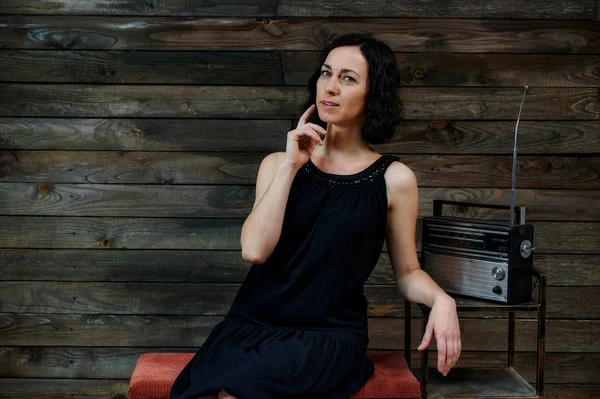 Retrato de una linda mujer morena emocional en un vestido negro sentado en una silla sobre un fondo gris alternativo en el interior vintage con una radio en un soporte . — Foto de Stock