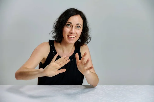 Portrait d'une jolie femme brune souriante parlant dans une robe noire sur un fond blanc. Assis à une table juste en face de la caméra avec des émotions vives . — Photo