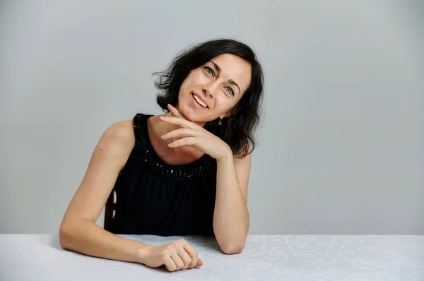 Portrait d'une jolie femme brune souriante parlant dans une robe noire sur un fond blanc. Assis à une table juste en face de la caméra avec des émotions vives . — Photo