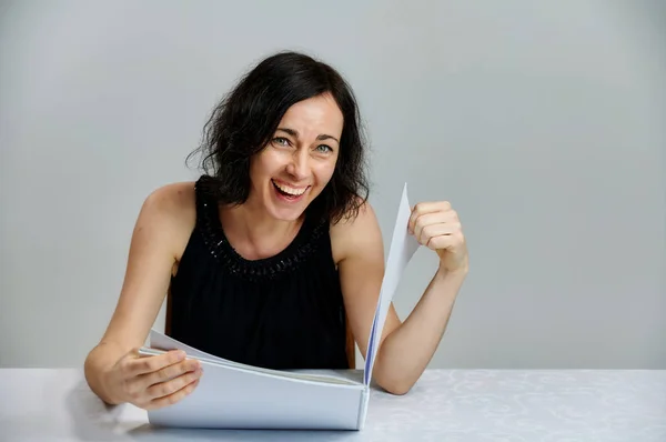 Portrait of a cute smiling talking brunette woman in a black dress on a white background. Sits at a table right in front of the camera with vivid emotions with a folder in his hands. — Stock Photo, Image