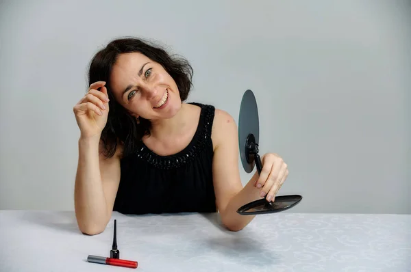 Portrait d'une jolie femme brune souriante parlant dans une robe noire sur un fond blanc. Assis à une table juste en face de la caméra avec des émotions vives avec un ensemble de cosmétiques . — Photo