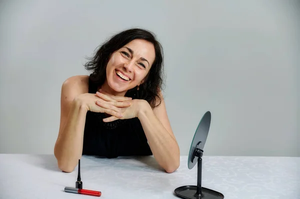 Portrait d'une jolie femme brune souriante parlant dans une robe noire sur un fond blanc. Assis à une table juste en face de la caméra avec des émotions vives avec un ensemble de cosmétiques . — Photo