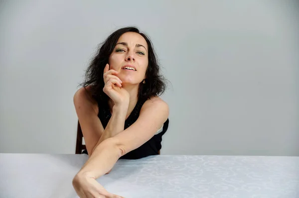 Modelo Se sienta en una mesa justo delante de la cámara con emociones vívidas. Retrato de una linda mujer morena sonriente con un vestido negro sobre un fondo blanco . — Foto de Stock