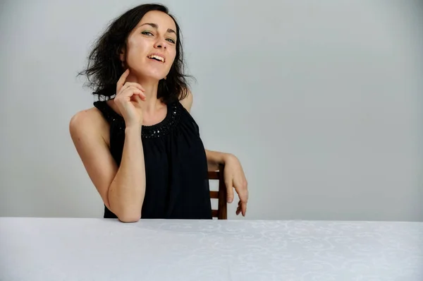 Modèle Assis à une table juste en face de la caméra avec des émotions vives. Portrait d'une jolie femme brune souriante parlant dans une robe noire sur un fond blanc . — Photo