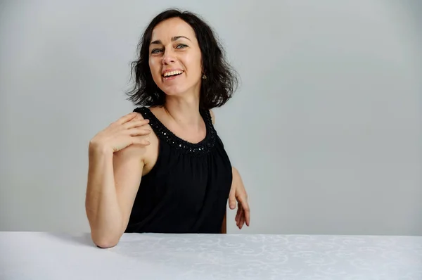 Modèle Assis à une table juste en face de la caméra avec des émotions vives. Portrait d'une jolie femme brune souriante parlant dans une robe noire sur un fond blanc . — Photo