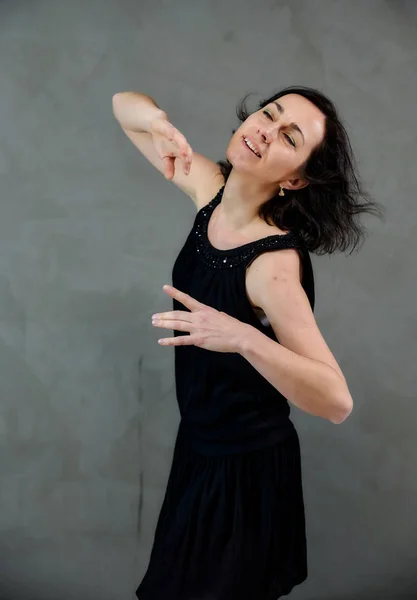 Modèle Debout juste devant la caméra avec des émotions vives. Portrait vertical d'une jolie femme brune souriante en robe noire sur fond gris alternatif . — Photo