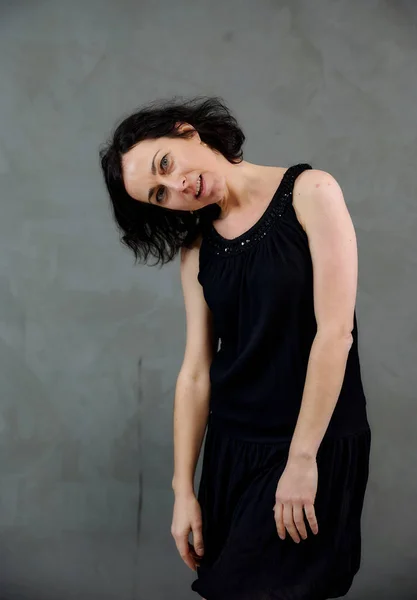 Modelo De pie justo delante de la cámara con emociones vívidas. Retrato vertical de una linda mujer morena sonriente en vestido negro sobre fondo alternativo gris . — Foto de Stock