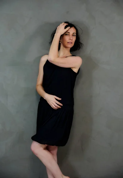 Model Standing right in front of the camera with vivid emotions. Vertical portrait of a cute smiling brunette woman in black dress on gray alternative background.