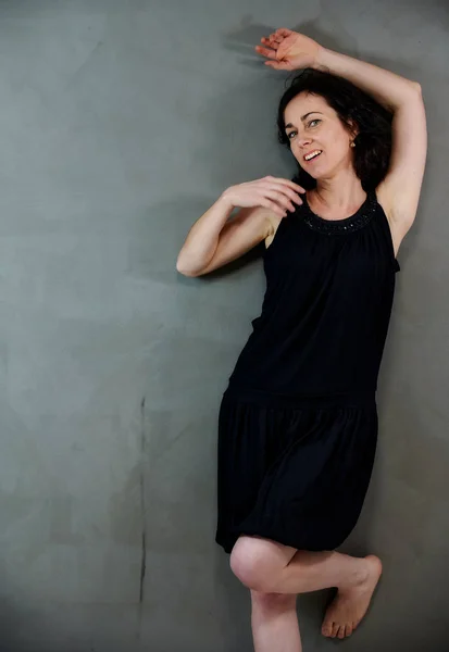 Modèle Debout juste devant la caméra avec des émotions vives. Portrait vertical d'une jolie femme brune souriante en robe noire sur fond gris alternatif . — Photo