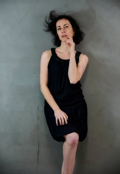 Modèle Debout juste devant la caméra avec des émotions vives. Portrait vertical d'une jolie femme brune souriante en robe noire sur fond gris alternatif . — Photo