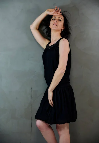 Modelo De pé bem na frente da câmera com emoções vivas. Retrato vertical de uma mulher morena sorridente bonito em vestido preto em fundo alternativo cinza . — Fotografia de Stock