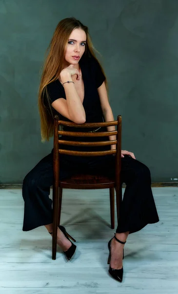 Het model zit in verschillende houdingen op een stoel voor de camera. Het concept van glamour, stijl. Portret van een mooi blond meisje met lang haar en uitstekende make-up op een grijze achtergrond. — Stockfoto
