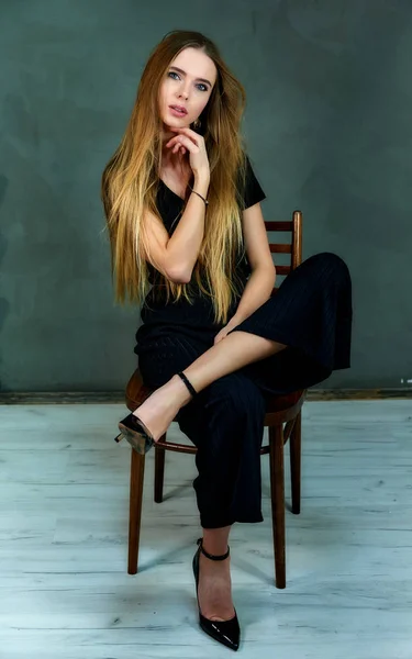 Το μοντέλο κάθεται σε διαφορετικές στάσεις σε μια καρέκλα μπροστά από την κάμερα. Η έννοια της αίγλης, στυλ. Πορτρέτο ενός όμορφου ξανθού κοριτσιού με μακριά μαλλιά και εξαιρετικό μακιγιάζ σε γκρι φόντο. — Φωτογραφία Αρχείου