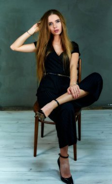 The model sits in different poses on a chair in front of the camera. The concept of glamor, style. Portrait of a pretty blonde girl with long hair and excellent make-up on a gray background.