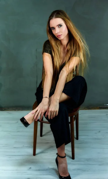 El modelo se sienta en diferentes poses en una silla delante de la cámara. El concepto de glamour, estilo. Retrato de una guapa rubia de pelo largo y excelente maquillaje sobre fondo gris . — Foto de Stock