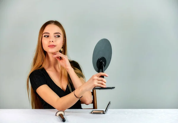 The model sits in different poses at the table and does makeup in front of the camera. Portrait of a pretty blonde girl with long hair and great makeup on a white background. — Stockfoto